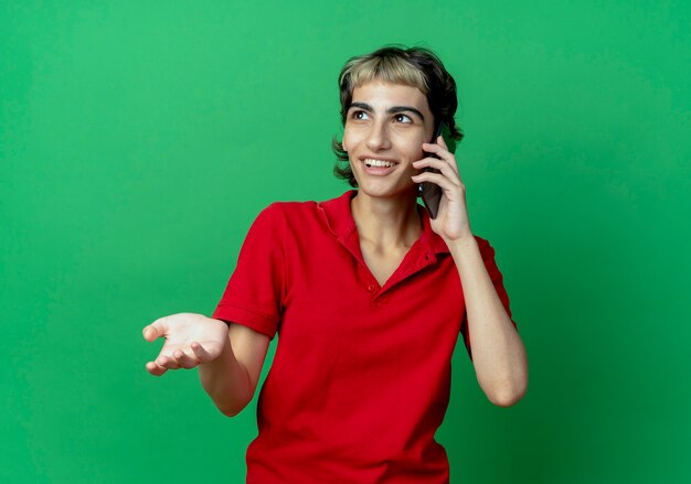 Souriante jeune fille de race blanche avec coupe de cheveux de lutin, parler au téléphone à côté isolé sur fond vert avec espace de copie