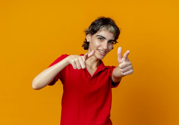 Souriante jeune fille de race blanche avec coupe de cheveux de lutin pointant vers la caméra isolée sur fond orange avec espace de copie