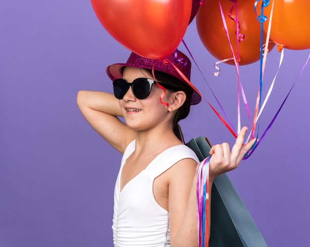 Souriante jeune fille de race blanche à lunettes de soleil avec chapeau de fête violet tenant des ballons d'hélium et des sacs à provisions sur l'épaule isolé sur un mur violet avec espace copie