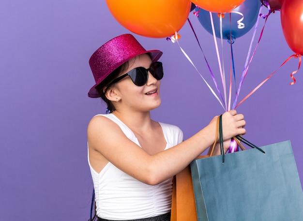 Souriante jeune fille de race blanche à lunettes de soleil avec chapeau de fête violet tenant des sacs à provisions et regardant des ballons d'hélium isolés sur un mur violet avec espace copie