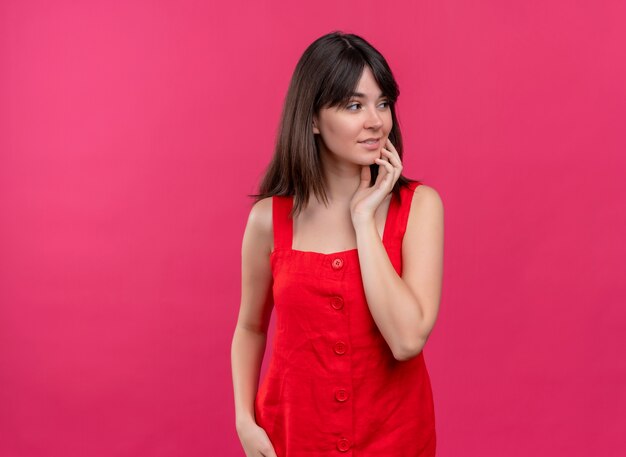 Souriante jeune fille de race blanche touchant le visage avec la main et regardant sur le côté sur fond rose isolé avec espace copie