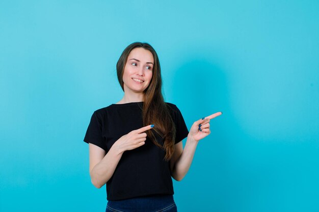 Souriante jeune fille regarde à droite en pointant vers la droite avec les index sur fond bleu