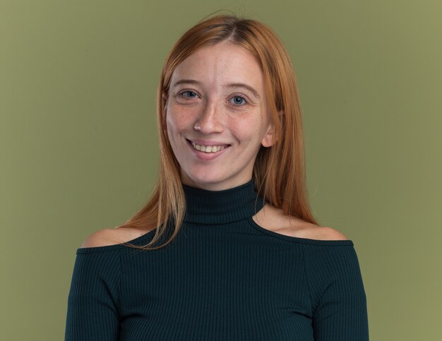 Souriante jeune fille rousse au gingembre avec des taches de rousseur isolée sur un mur vert olive avec espace de copie