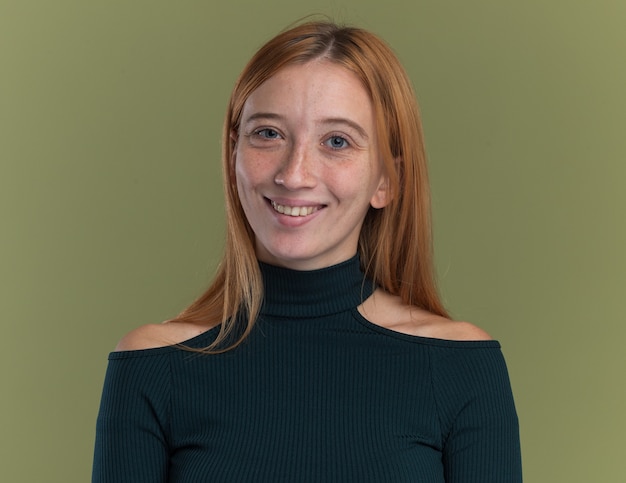 Souriante jeune fille rousse au gingembre avec des taches de rousseur isolée sur un mur vert olive avec espace de copie