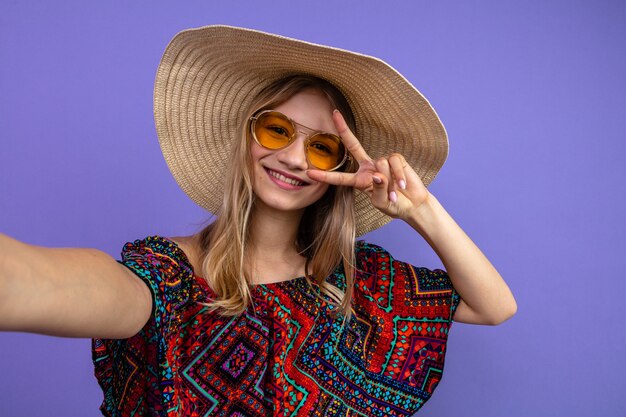 Souriante jeune fille slave blonde avec des lunettes de soleil et avec un chapeau de soleil gesticulant le signe de la victoire et