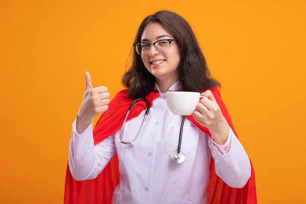 Souriante jeune fille de super-héros caucasien portant un uniforme de médecin et un stéthoscope avec des lunettes tenant une tasse de thé montrant le pouce vers le haut isolé sur le mur