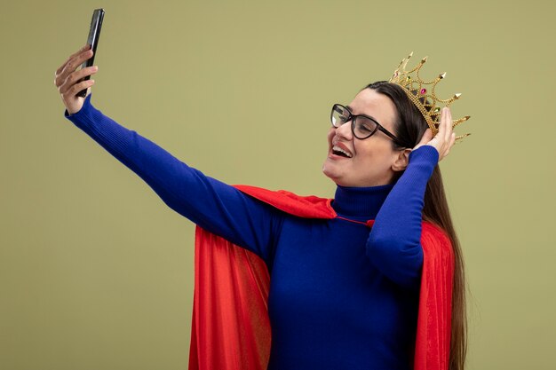 Souriante jeune fille de super-héros portant des lunettes et une couronne mettant la main sur la couronne et prendre selfie isolé sur fond vert olive