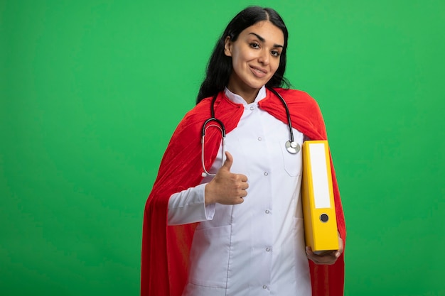 Photo gratuite souriante jeune fille de super-héros portant une robe médicale avec stéthoscope tenant le dossier montrant le pouce vers le haut isolé sur vert