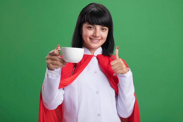 Photo gratuite souriante jeune fille de super-héros portant un stéthoscope avec une robe médicale et une cape tenant une tasse de thé montrant le pouce vers le haut isolé sur vert