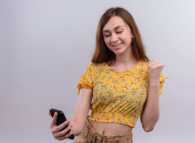 Souriante jeune fille tenant un téléphone mobile avec le poing levé sur un mur blanc isolé