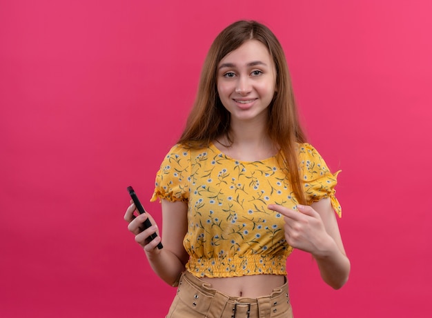 Souriante jeune fille tenant un téléphone mobile et pointant dessus sur un mur rose isolé avec espace de copie