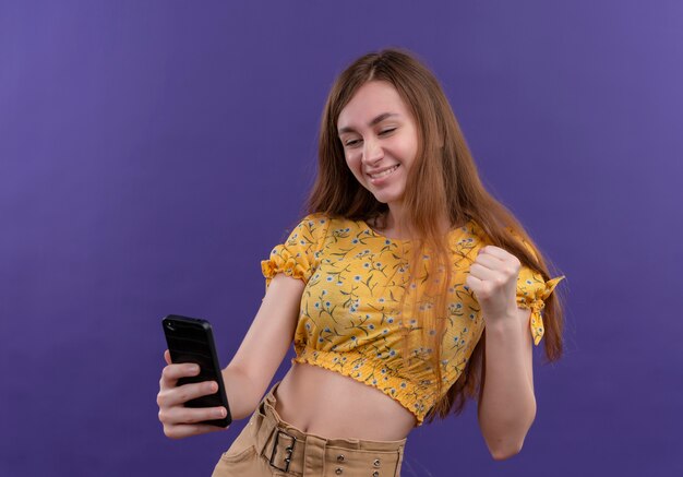 Souriante jeune fille tenant un téléphone portable et levant le poing sur un mur violet isolé