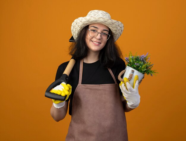 Souriante jeune jardinière brune à lunettes optiques et en uniforme portant un chapeau et des gants de jardinage détient pot de fleurs et pelle sur l'épaule isolé sur un mur orange avec espace de copie