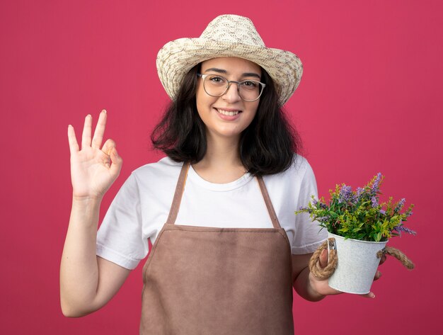 Souriante jeune jardinière brune à lunettes optiques et en uniforme portant un chapeau de jardinage détient pot de fleurs et gestes ok signe de la main isolé sur un mur rose avec espace de copie