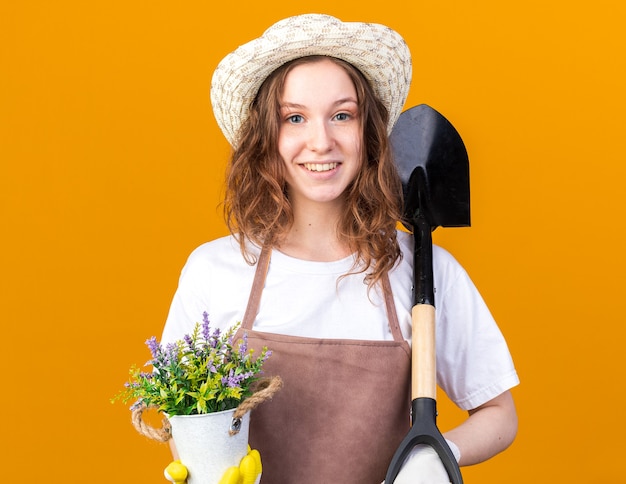 Souriante Jeune Jardinière Portant Un Chapeau De Jardinage Avec Des Gants Tenant Une Fleur Dans Un Pot De Fleurs Avec Une Pelle Isolée Sur Un Mur Orange