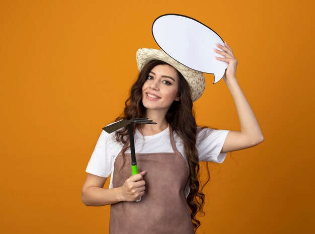 Souriante jeune jardinière en uniforme portant chapeau de jardinage détient bulle et houe râteau isolé sur mur orange avec espace de copie