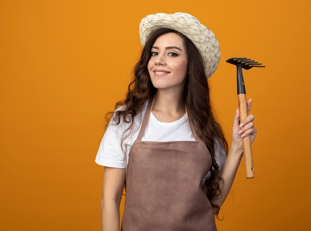 Souriante jeune jardinière en uniforme portant chapeau de jardinage détient râteau isolé sur mur orange avec espace copie