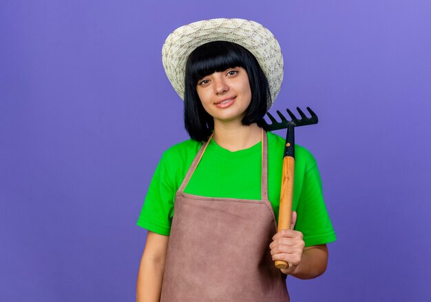 Souriante jeune jardinière en uniforme portant un chapeau de jardinage détient un râteau
