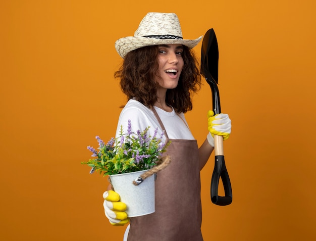 Souriante Jeune Jardinière En Uniforme Portant Un Chapeau De Jardinage Et Des Gants Tenant Des Fleurs En Pot De Fleurs