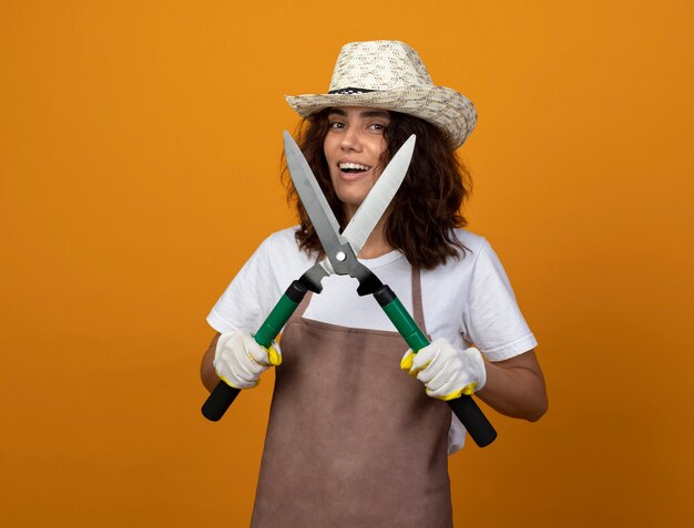 Souriante jeune jardinière en uniforme portant un chapeau de jardinage et des gants tenant une tondeuse