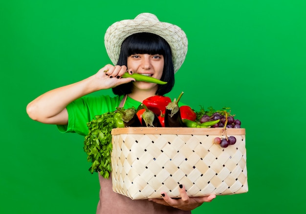 Photo gratuite souriante jeune jardinière en uniforme portant un chapeau de jardinage tenant un panier de légumes