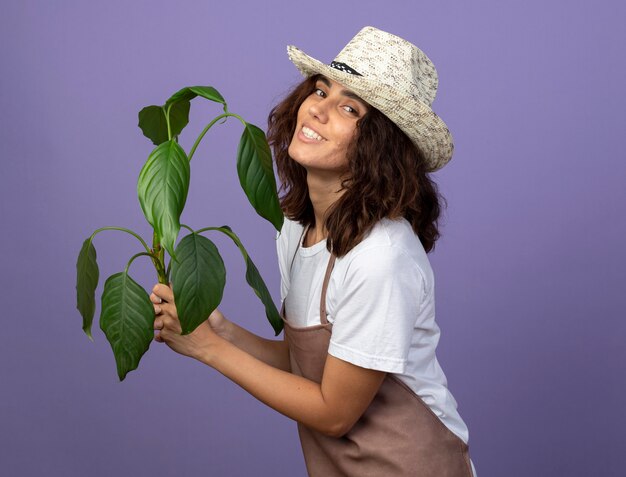 Souriante jeune jardinière en uniforme portant un chapeau de jardinage tenant une plante