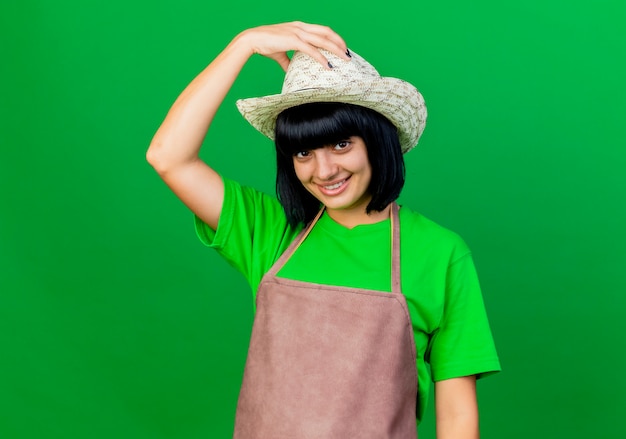 Souriante jeune jardinière en uniforme portant et tenant un chapeau de jardinage