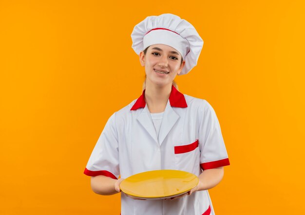 Souriante jeune jolie cuisinière en uniforme de chef avec appareil dentaire tenant une assiette vide isolée sur l'espace orange