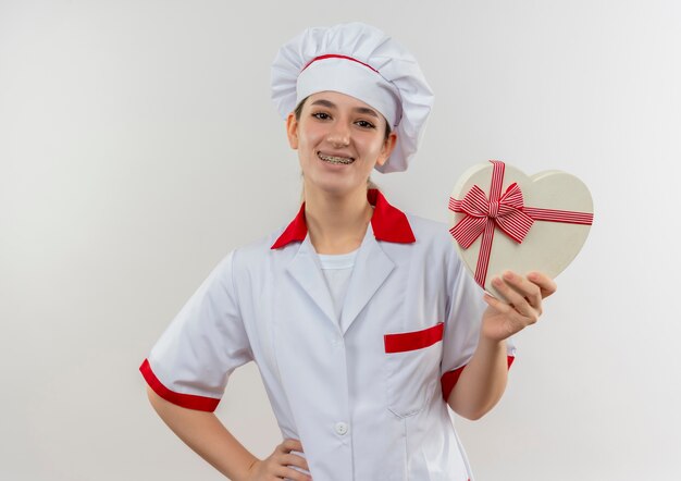 Souriante jeune jolie cuisinière en uniforme de chef avec appareil dentaire tenant une boîte-cadeau en forme de coeur avec la main sur la taille isolée sur l'espace blanc