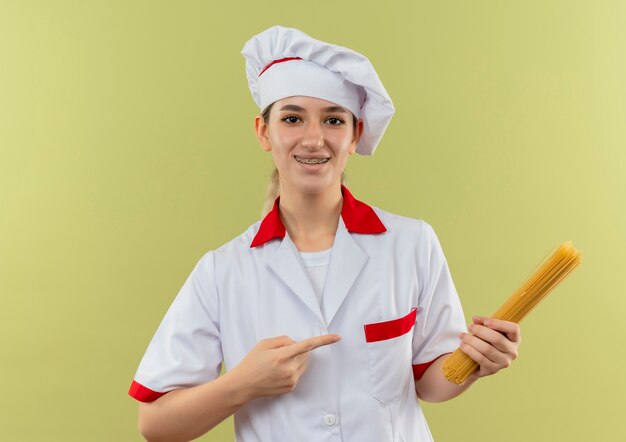 Souriante jeune jolie cuisinière en uniforme de chef avec appareil dentaire tenant et pointant sur les pâtes spaghetti isolé sur l'espace vert