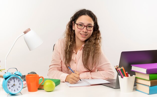 Souriante jeune jolie écolière portant des lunettes assis au bureau avec des outils scolaires à faire ses devoirs en écrivant sur le bloc-notes isolé sur un mur blanc