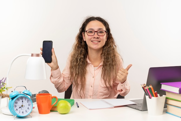 Souriante jeune jolie écolière portant des lunettes assis au bureau avec des outils scolaires à faire ses devoirs montrant un téléphone mobile et le pouce vers le haut isolé sur un mur blanc