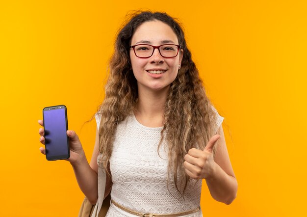 Souriante jeune jolie écolière portant des lunettes et sac à dos montrant le téléphone mobile et le pouce vers le haut isolé sur mur jaune