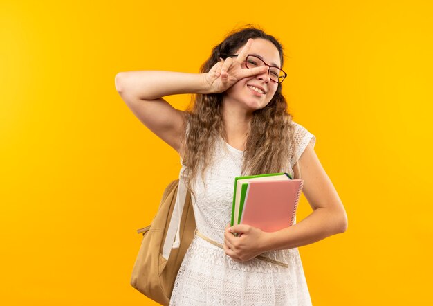 Souriante jeune jolie écolière portant des lunettes et sac à dos tenant livre et bloc-notes faisant signe de paix isolé sur mur jaune