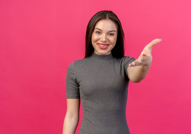 Souriante jeune jolie femme étendant la main à la caméra isolée sur rose avec espace copie