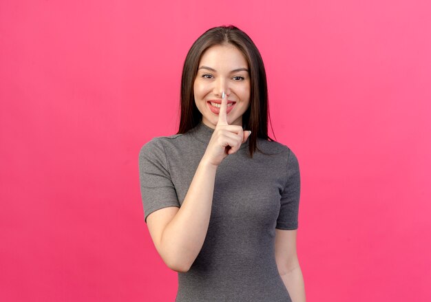 Souriante jeune jolie femme faisant le geste de silence isolé sur fond rose avec espace copie