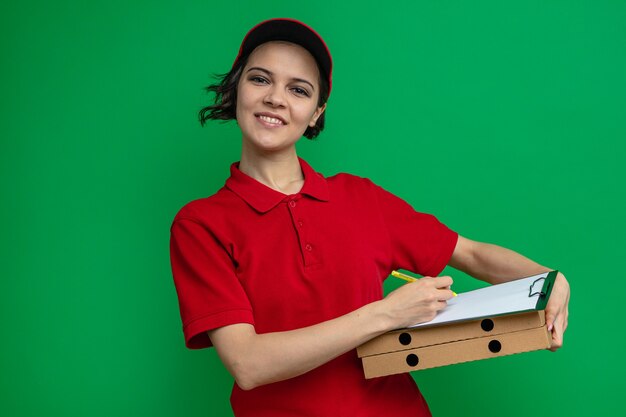 Souriante jeune jolie femme de livraison tenant un presse-papiers sur des boîtes à pizza