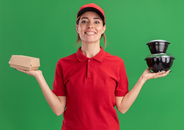 Souriante jeune jolie femme de livraison en uniforme détient des contenants de nourriture et un paquet de nourriture à l'avant isolé sur mur vert