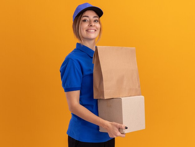 Souriante jeune jolie femme de livraison en uniforme détient un paquet de papier sur carton isolé sur mur orange