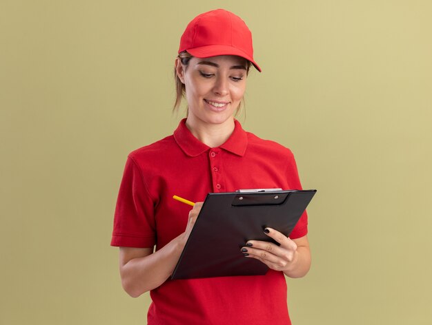 Souriante jeune jolie femme de livraison en uniforme écrit sur le presse-papiers avec un stylo isolé sur mur vert olive