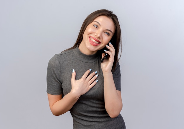 Souriante jeune jolie femme parlant au téléphone à la recherche de mettre la main droite sur la poitrine isolé sur fond blanc avec espace copie