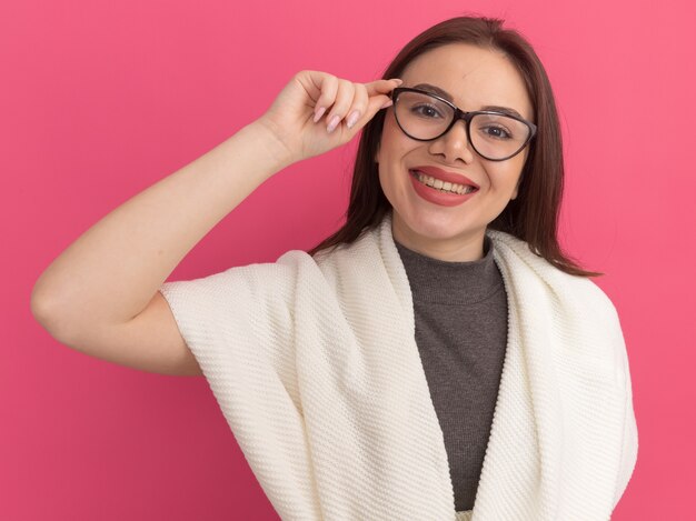 Souriante jeune jolie femme portant et saisissant des lunettes isolées sur un mur rose