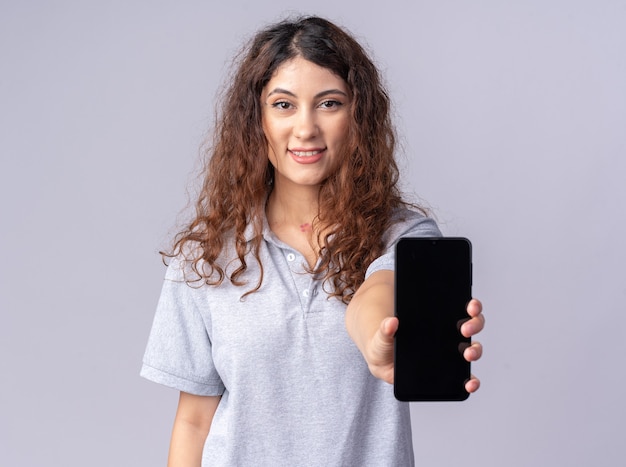 Souriante jeune jolie femme regardant devant étirant le téléphone portable vers l'avant isolé sur mur blanc