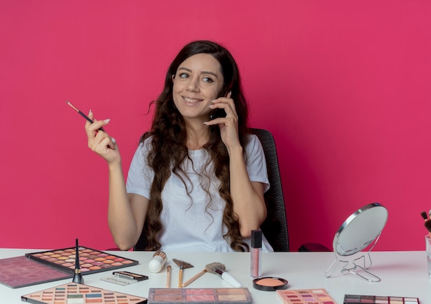 Photo gratuite souriante jeune jolie fille assise à la table de maquillage avec des outils de maquillage parlant au téléphone et tenant une brosse de fard à paupières regardant côté isolé sur fond cramoisi