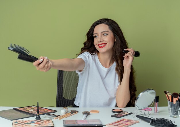 Souriante jeune jolie fille assise à la table de maquillage avec des outils de maquillage tenant et étirant des peignes et regardant à côté isolé sur fond vert olive