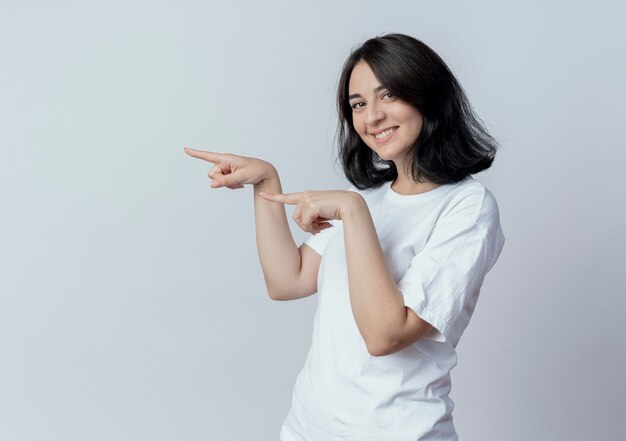 Souriante jeune jolie fille caucasienne debout en vue de profil et pointant tout droit isolé sur fond blanc avec espace de copie