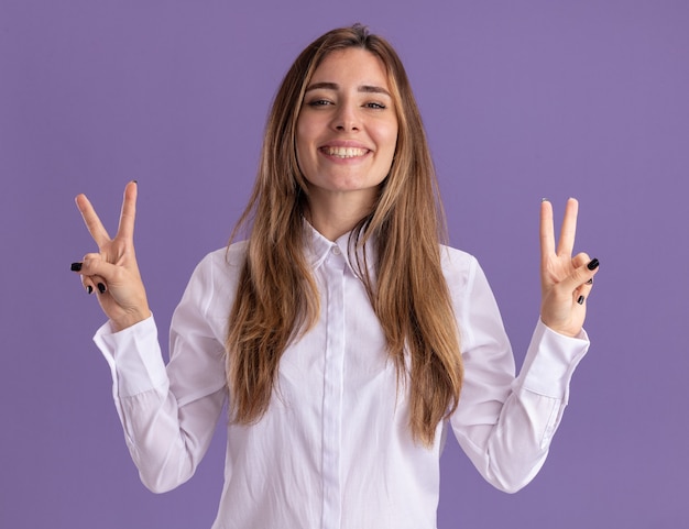 Souriante jeune jolie fille caucasienne gestes signe de victoire avec deux mains sur violet