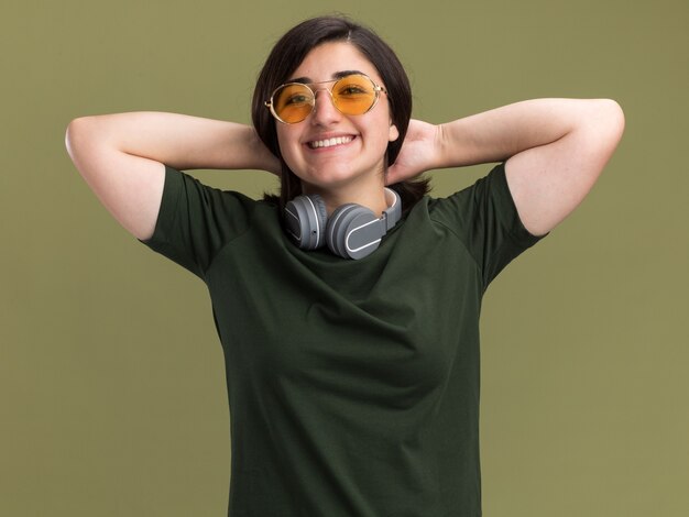 Souriante jeune jolie fille caucasienne à lunettes de soleil et avec des écouteurs autour du cou met les mains sur le cou derrière isolé sur un mur vert olive avec espace de copie