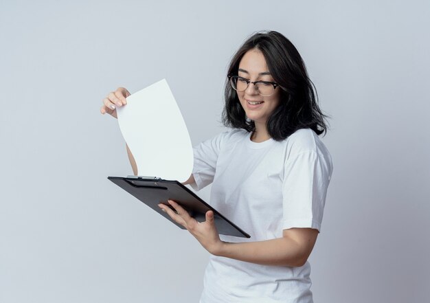 Souriante jeune jolie fille caucasienne portant des lunettes tenant et regardant le presse-papiers isolé sur fond blanc