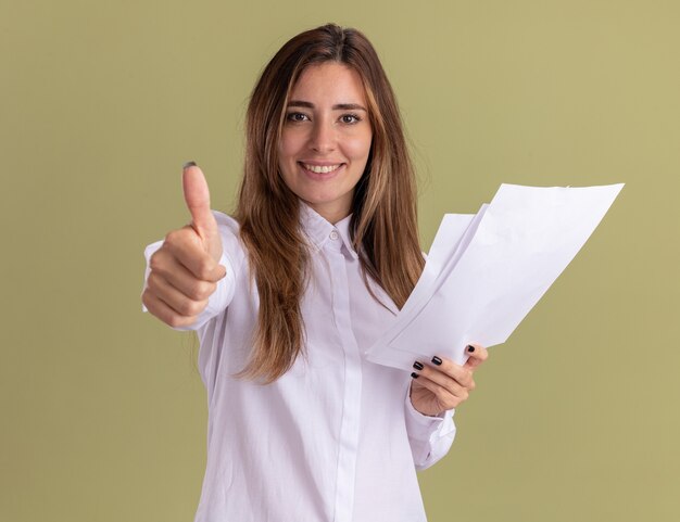 Souriante jeune jolie fille caucasienne tient des feuilles de papier vierges et les pouces vers le haut isolés sur un mur vert olive avec espace de copie